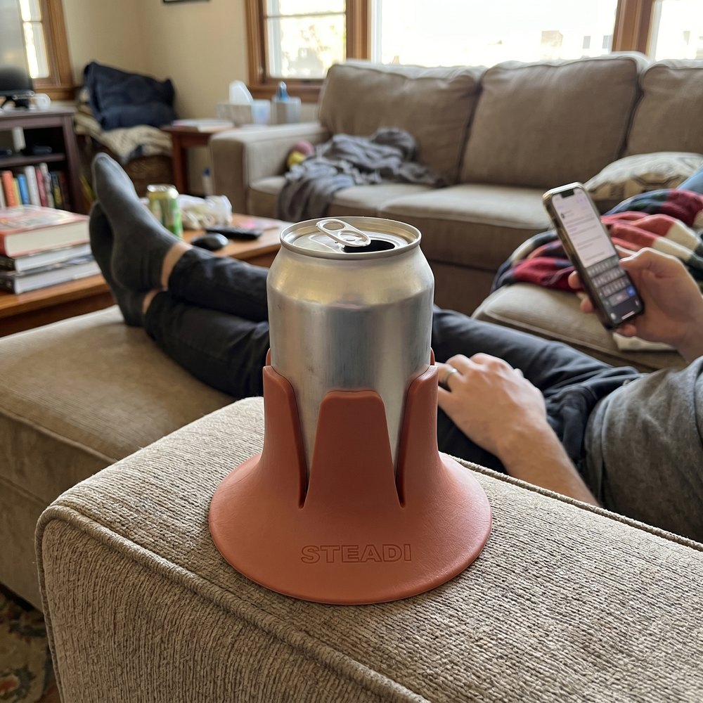 A can standing in a terracotta Steadi on a sofa cushion beside someone reclined with their feet up.